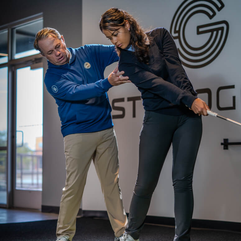 Golf coach assisting a player with swing technique during an indoor lesson at Studio Golf San Diego.