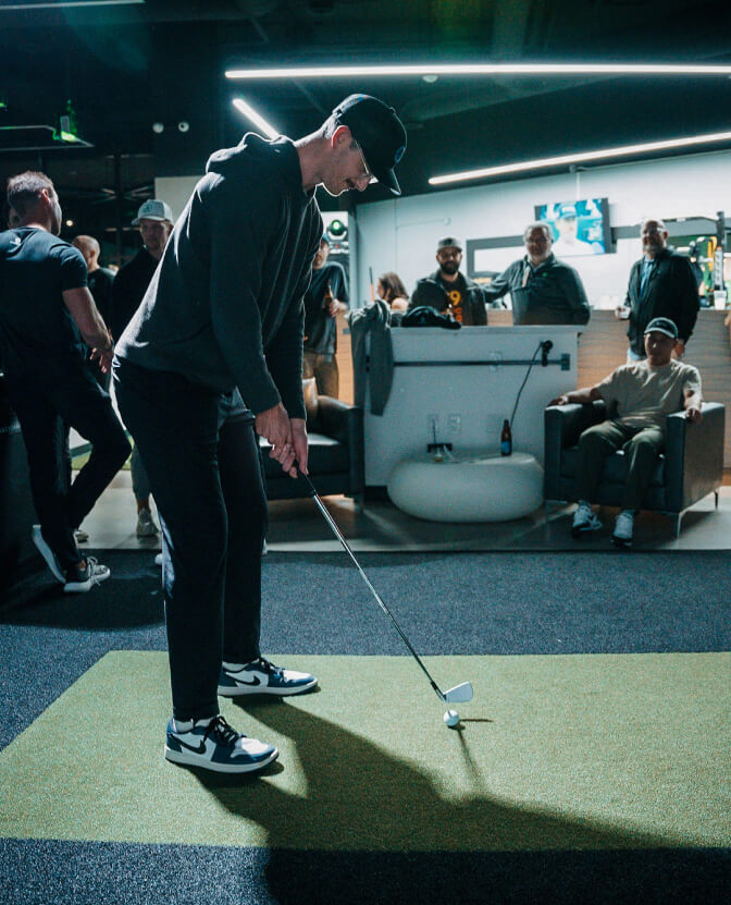 Golfer practicing in a premium hitting bay at Studio Golf while other members watch in a modern training lounge.